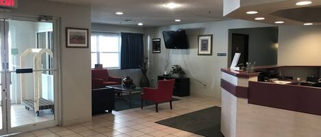 A hotel lobby with a reception desk, a TV mounted on the wall, a red chair, a coffee table, and a window with curtains.