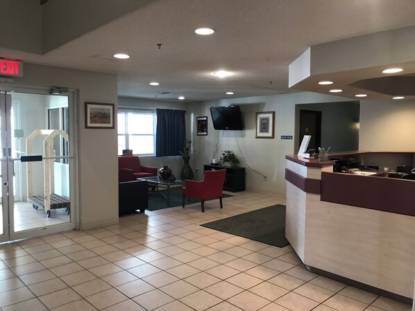 A hotel lobby with a reception desk, a TV mounted on the wall, a red chair, a coffee table, and a window with curtains. - Chadron Inn & Suites (Chadron)
