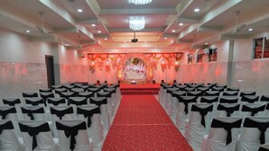 A banquet hall with rows of white chairs with black sashes, a red carpet aisle, and a stage with floral decorations. - Metropolis Hotel (Hubli)