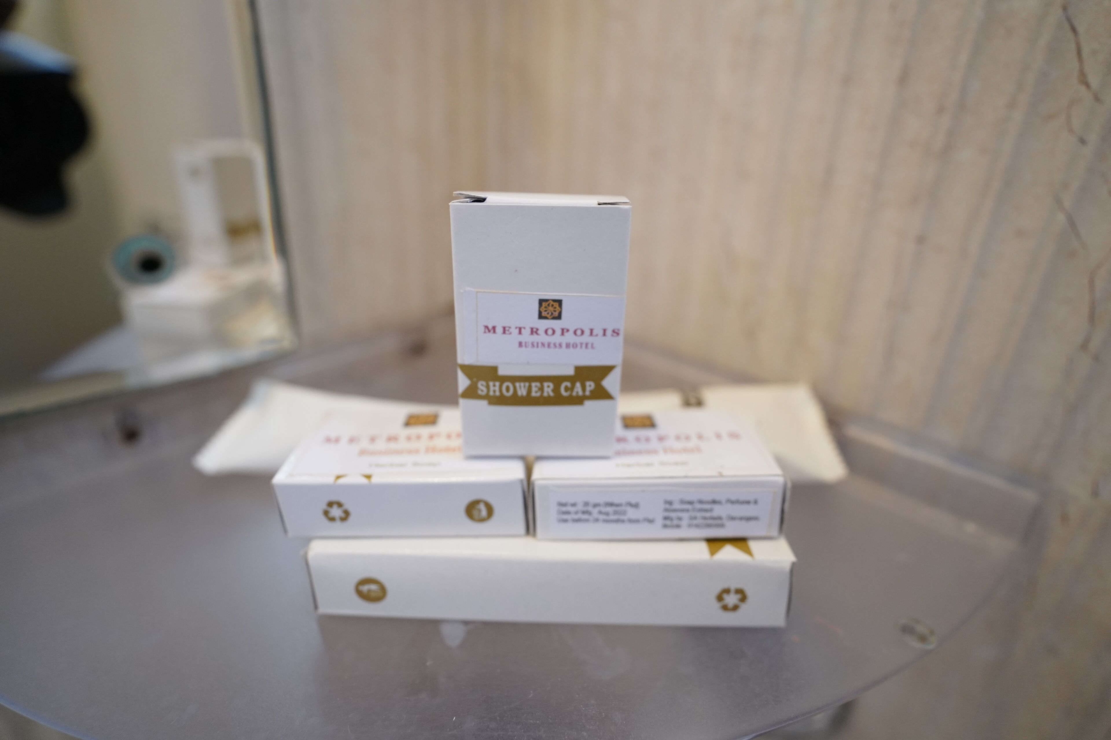 Un baño con espejo, lavamanos y una caja de gorro de ducha con la etiqueta 'Metropolis Business Hotel'.
