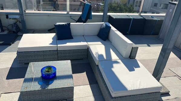 Una terraza en la azotea con un sofá modular, una pequeña mesa con una planta y una silla azul.