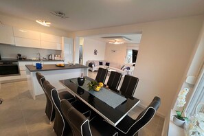 Una cocina moderna con armarios blancos, un comedor con mesa de vidrio y sillas negras, y una sala de estar con sofá y televisor.