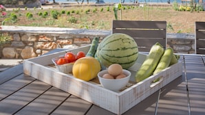 A wooden table with a fruit and egg tray, overlooking a landscaped garden and a body of water. - SEASIDE VILLA MARIA (PAROS)