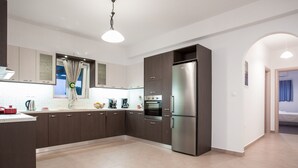 A modern kitchen with dark cabinets, a stainless steel refrigerator, and a large window with curtains. - SEASIDE VILLA MARIA (PAROS)