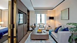 Une chambre d’hôtel moderne avec un grand lit, un canapé, une table basse avec des fruits, une télévision et une vue sur la ville.