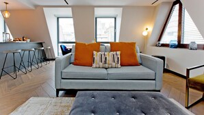 Ein modernes Wohnzimmer mit einem grauen Sofa, orangefarbenen Kissen und einem grauen Hocker. Zwei Fenster mit Jalousien, eine Küchenzeile mit Barhocker und eine Pendelleuchte sind ebenfalls vorhanden.
