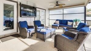 A sunlit outdoor seating area with wicker furniture and blue cushions, a ceiling fan, and a large window providing a view of the outdoors. - Luxe Jupiter Home w/ Pool House, Near Golf & Beach (Jupiter)