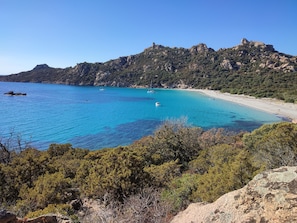 A coastal landscape with a clear blue sea, a sandy beach, and a rocky mountain backdrop.