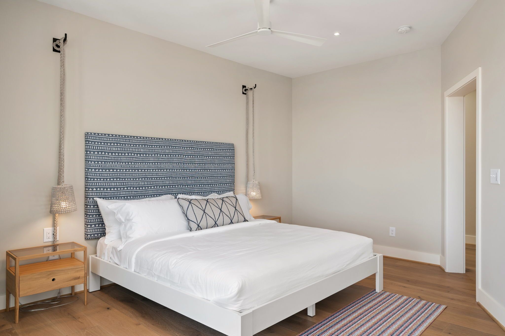 Ein Schlafzimmer mit einem weißen Bett, einem gemusterten Kopfteil, einem kleinen Nachttisch und einem Deckenventilator.