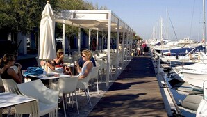 An outdoor dining area with white tables and chairs, a covered walkway, and boats docked in the background.