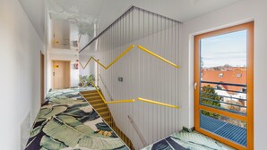 Un escalier avec des rampes jaunes, une moquette à motifs et une vue sur des immeubles par une fenêtre.