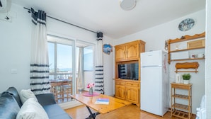 A compact living space with a sofa, a dining table, a kitchen area with wooden cabinets and a refrigerator, and a balcony with a view.