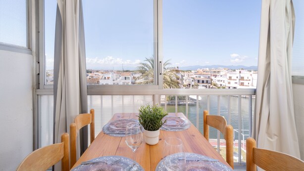 A balcony with a wooden table set for dining, a potted plant, and a view of a coastal town.