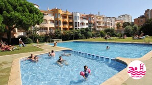 A swimming pool surrounded by multi-story buildings with balconies, a grassy area, and people swimming and relaxing.