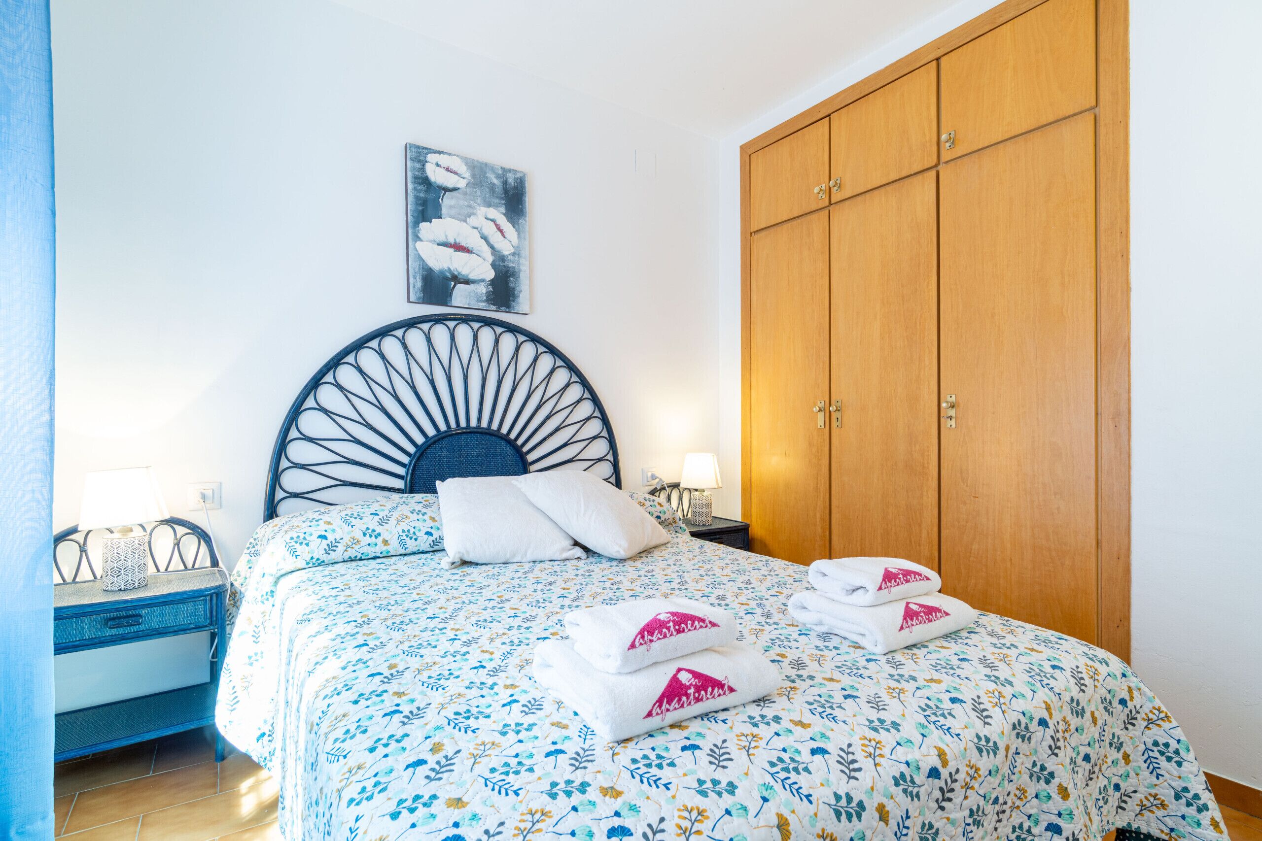 Een net opgemaakt bed met bloemenbeddels, een houten garderobe en een kunstwerk aan de muur.