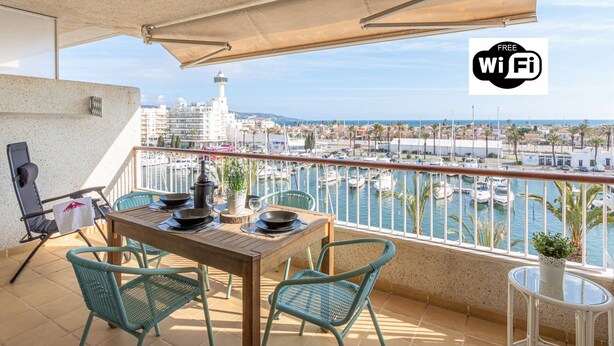 Ein Balkon mit Holztisch, zwei Stühlen, einem Blumentopf und Blick auf einen Yachthafen mit Booten.