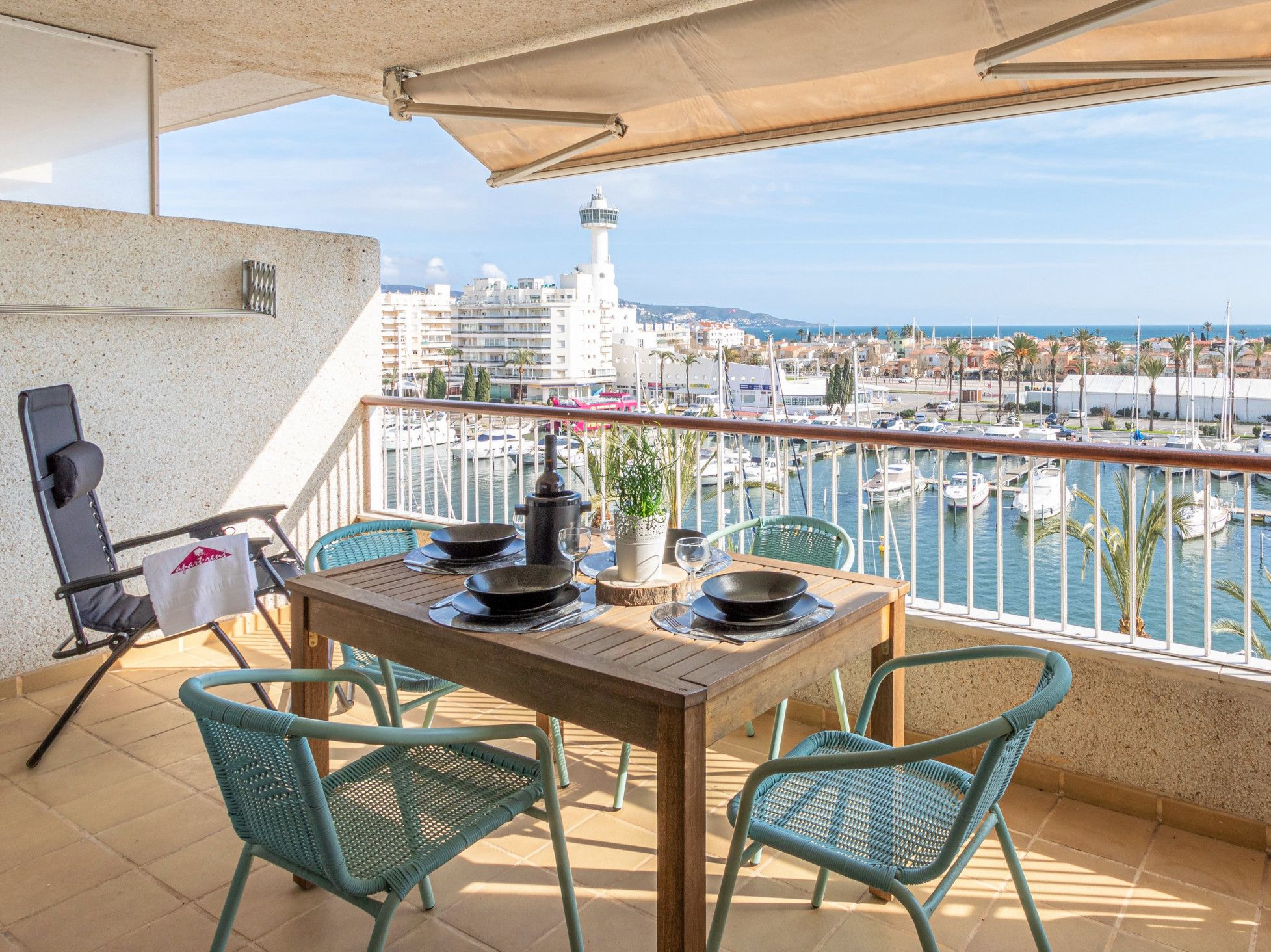 Een balkon met een houten tafel, twee stoelen, een bloempot en uitzicht op een jachthaven met boten.