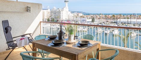 Un balcón con mesa de madera, dos sillas, una planta en maceta y vista a un puerto deportivo con barcos.