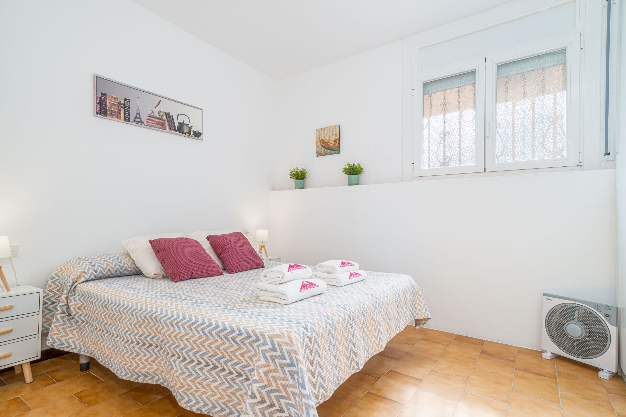 Un dormitorio con cama, almohadas, una mesita de noche, estante en la pared con plantas, ventana con cortinas y un ventilador de pie.