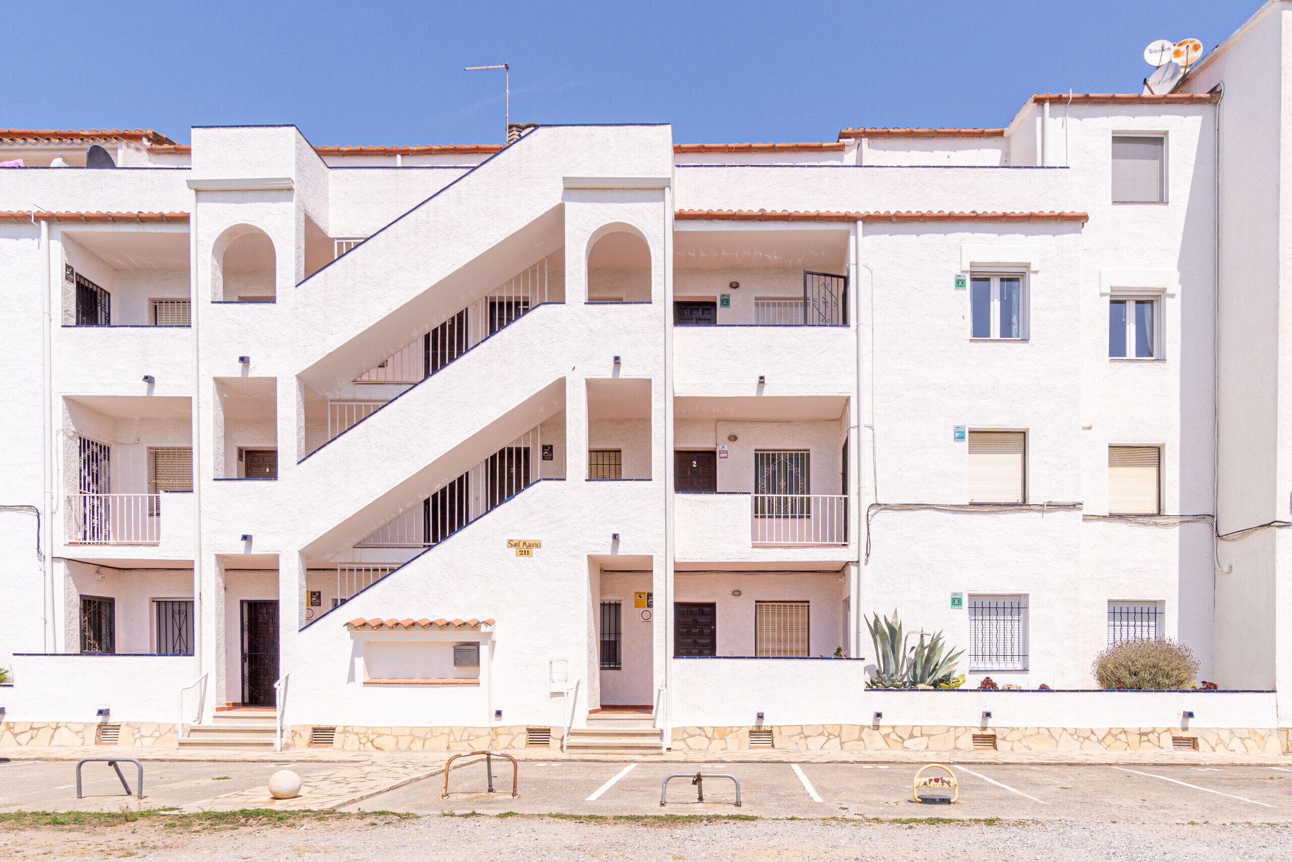 Un immeuble blanc à plusieurs étages, doté de balcons, d’un escalier central et d’un espace de stationnement à l’avant.