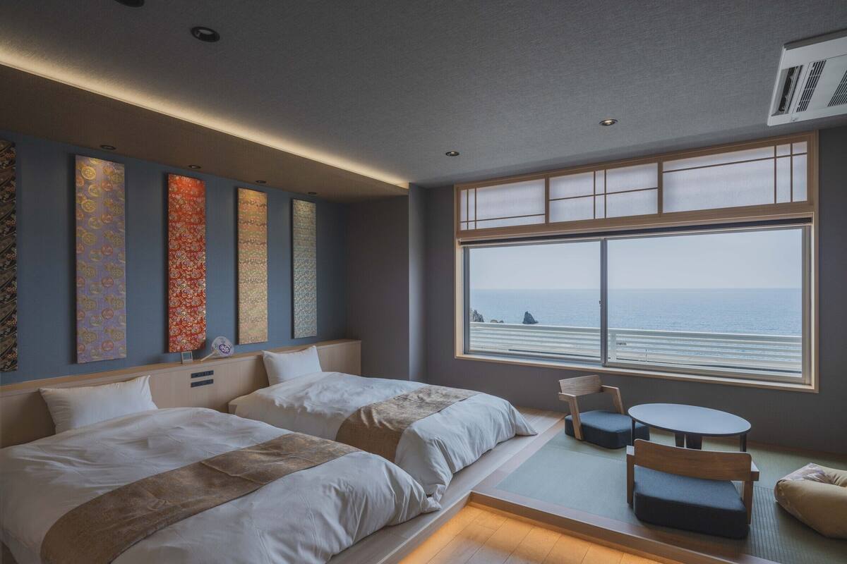 Ein Hotelzimmer mit großem Fenster, zwei Betten, einem kleinen Tisch und einem Sessel.