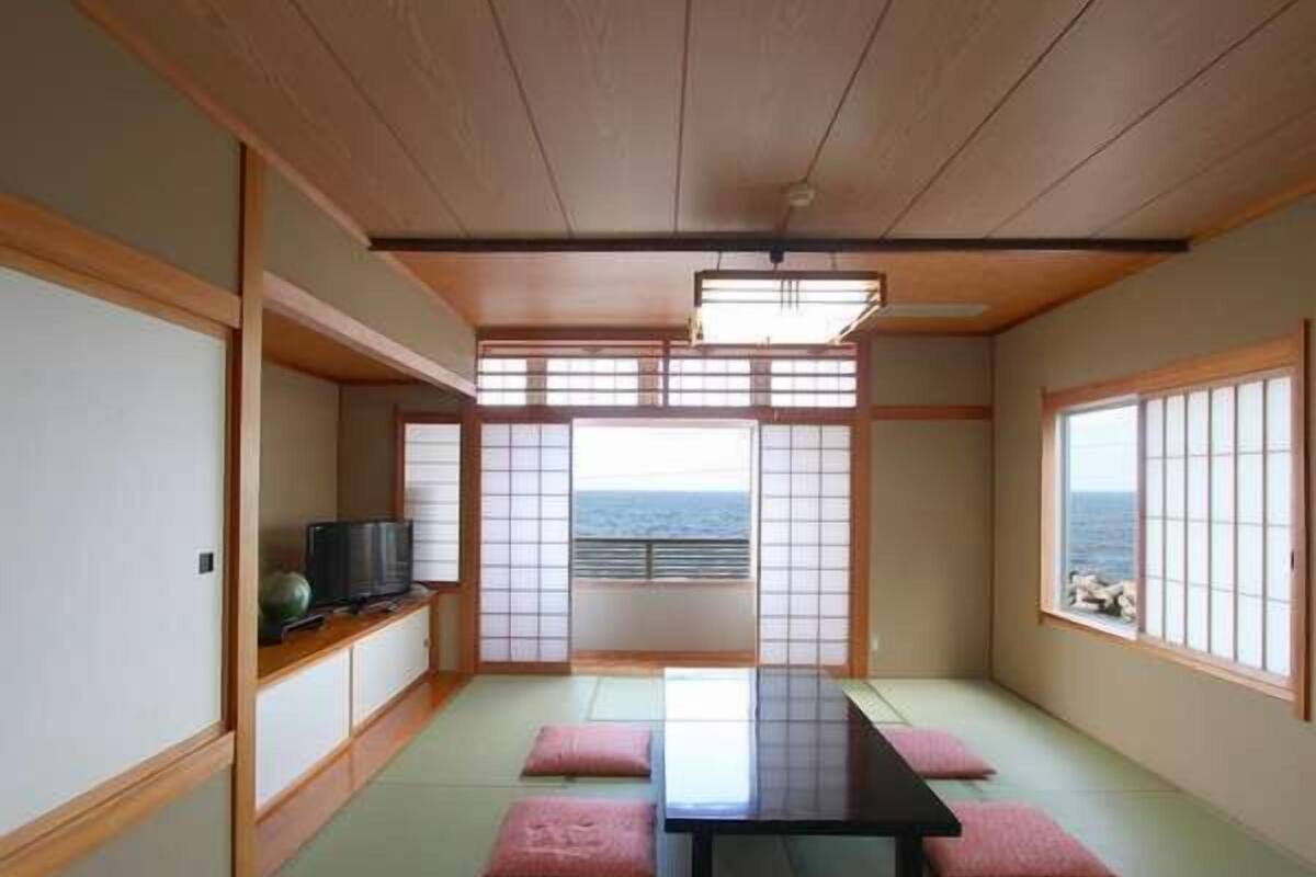 Ein traditioneller japanischer Raum mit Tatami-Boden, einem niedrigen Tisch, Kissen, Schiebetüren und Blick auf das Meer.