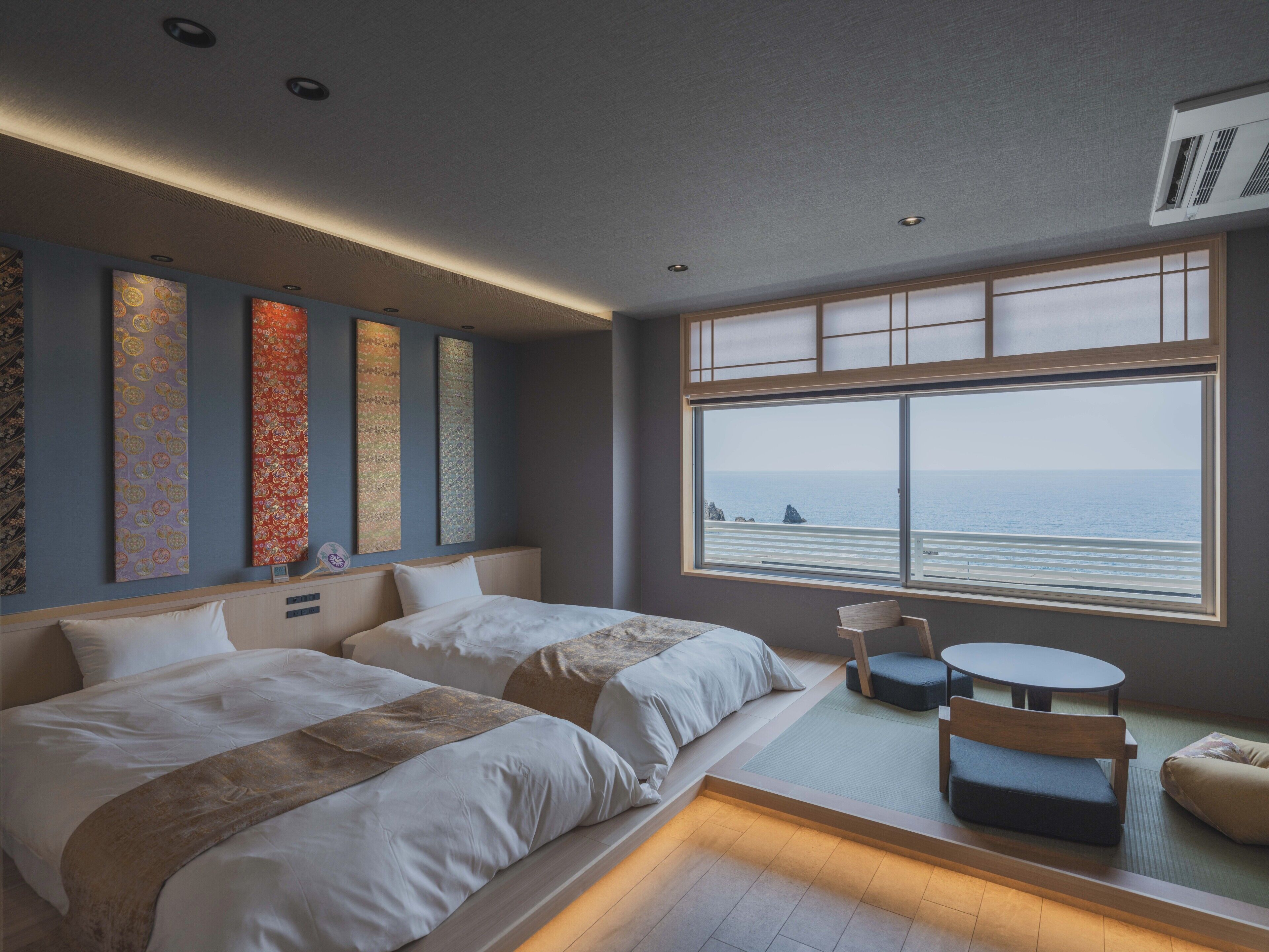 Een hotelkamer met twee bedden, een tafeltje, een stoel en een groot raam met uitzicht op zee.