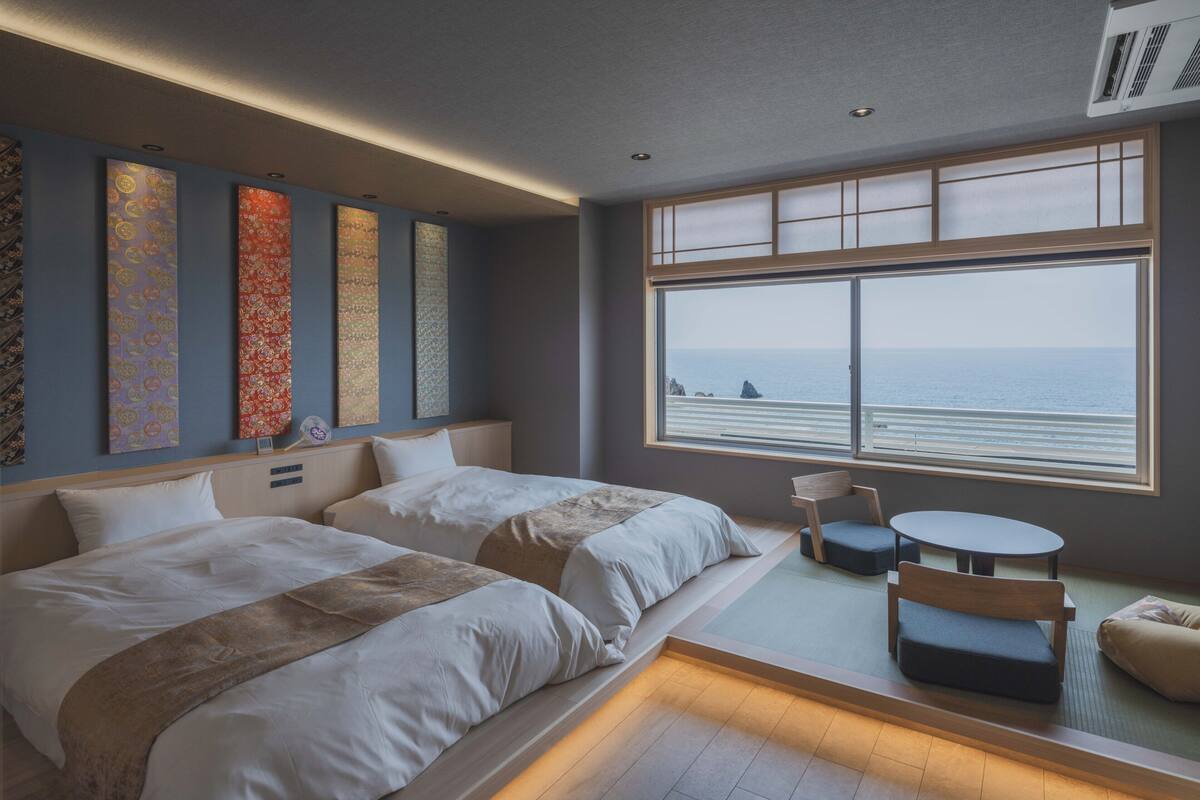 Ein Hotelzimmer mit zwei Betten, einem kleinen Tisch, einem Sessel und einem großen Fenster mit Meerblick.