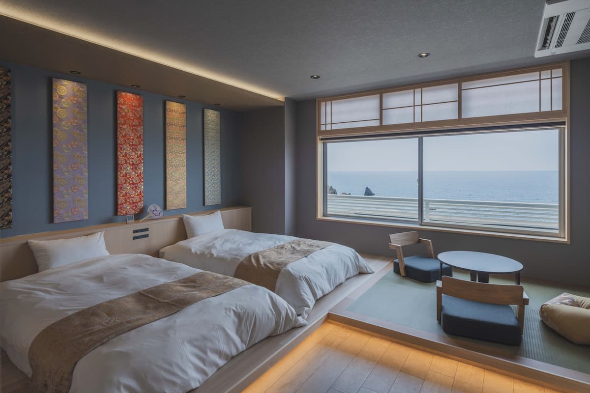 A hotel room with two beds, a small table, a chair, and a large window with a view of the sea.