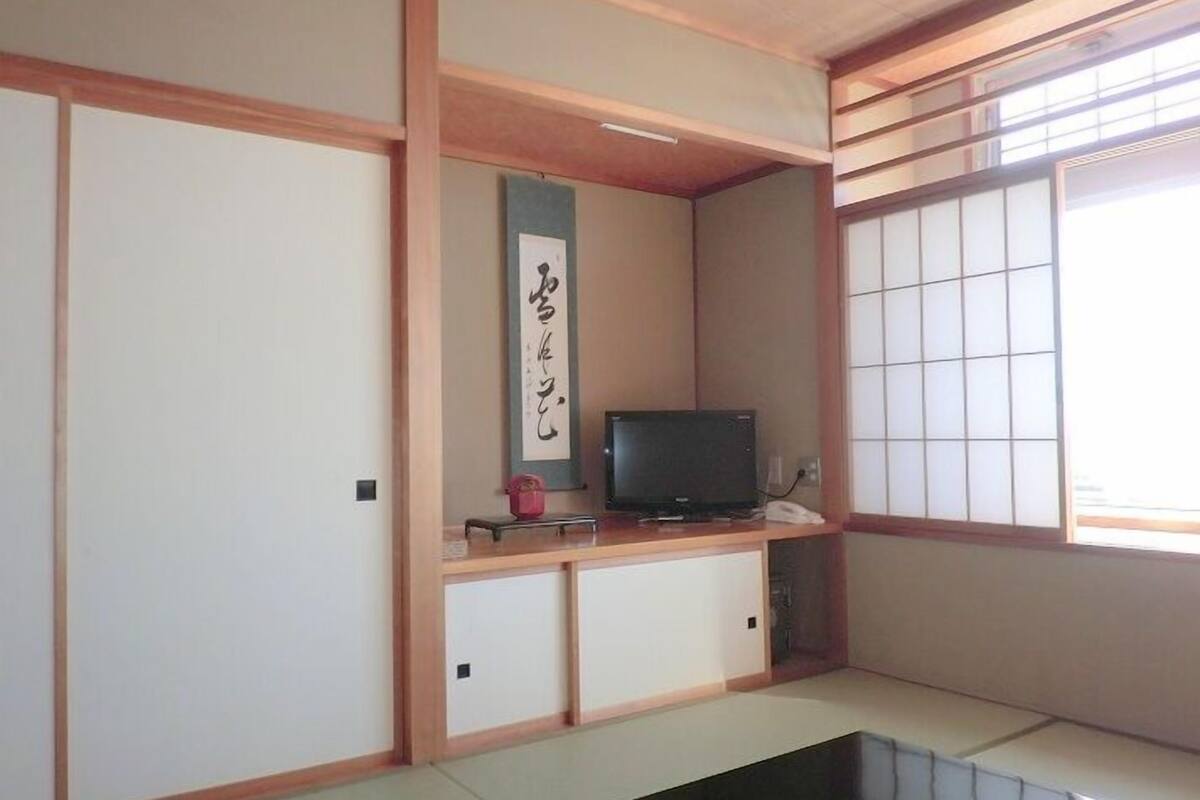 Ein traditioneller japanischer Raum mit Tatami-Boden, einer Schiebetür, einem Fernseher auf einer niedrigen Ablage und einem Shoji-Bildschirm.