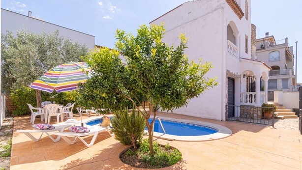A pool area with lounge chairs and a striped umbrella, surrounded by greenery and a white building with a tiled roof. - 0191-SANT MAURICI House with private pool (Empuriabrava)