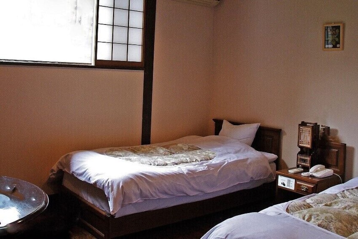 A bedroom with two beds, a bedside table with a phone, and a window with blinds.