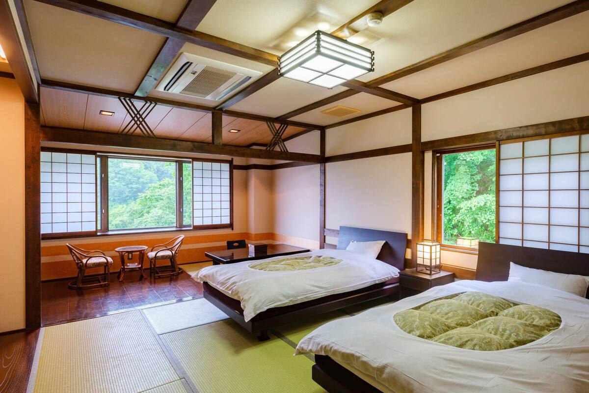 A traditional Japanese-style room with tatami flooring, two beds with green bedding, a wooden ceiling, and large windows with a view of greenery.