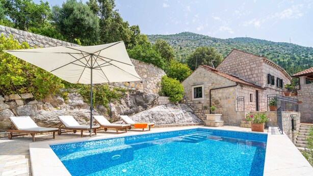 Une piscine entourée de bâtiments en pierre et de verdure, avec des chaises longues et un parasol.