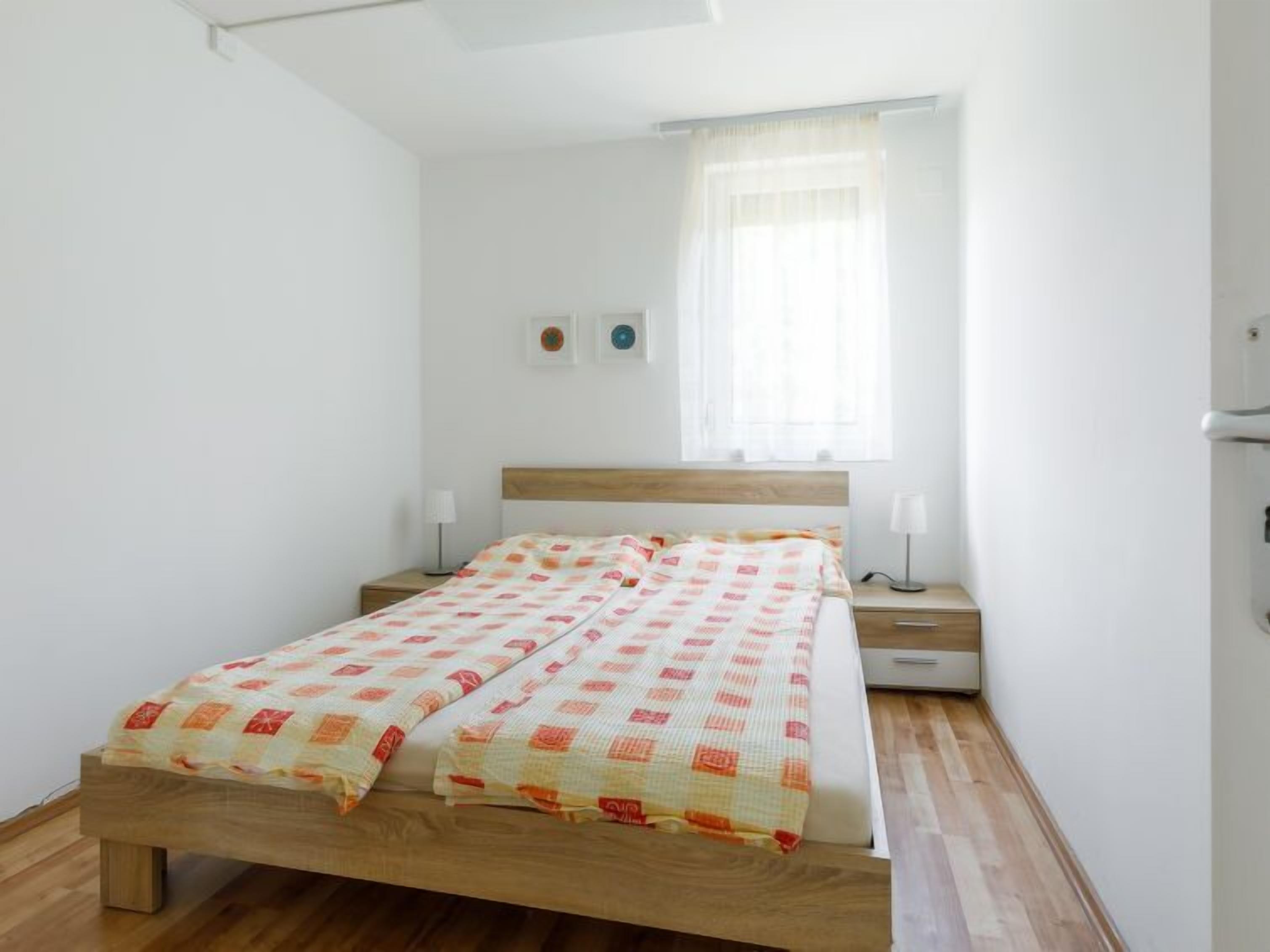 Ein Einzelbett mit Holzrahmen und einem gemusterten Bettüberwurf, zwei Nachttische mit Lampen, ein Fenster mit durchsichtigen Vorhängen sowie ein an der Wand befestigter Lichtschalter.
