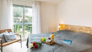 Un dormitorio con una cama grande, una silla de mimbre, una bandeja de desayuno con frutas y una botella, y vistas a la vegetación exterior.