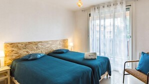 Habitación de hotel con dos camas, una banca, una lámpara y vistas al exterior a través de cortinas traslúcidas.