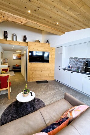 A modern living room with a sofa, a TV, and a kitchen area with white cabinets and a stone backsplash.