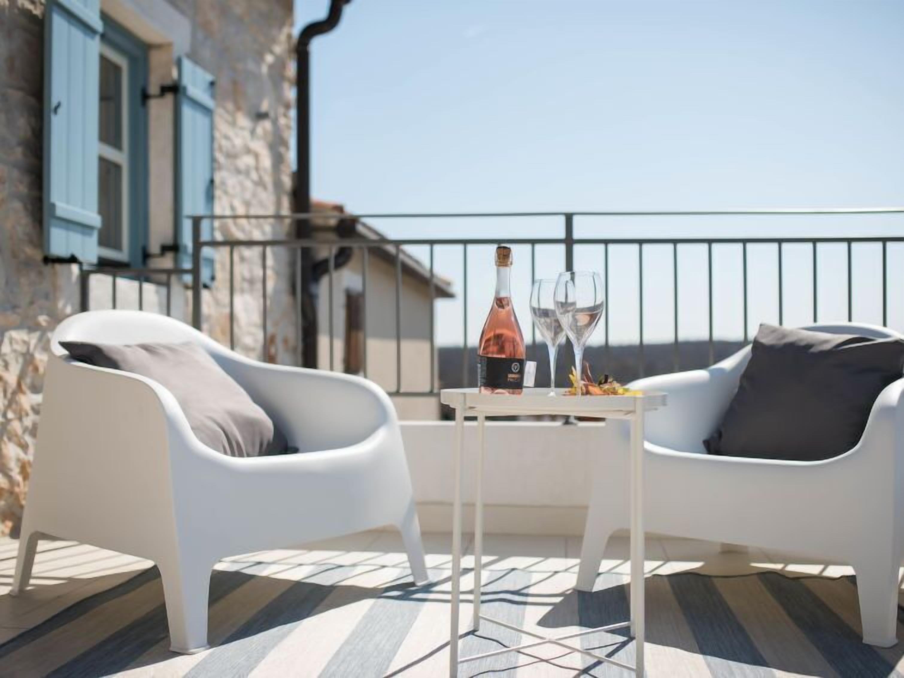 Ein Balkon mit zwei weißen Stühlen, einem kleinen Tisch mit Flasche und Gläsern, und einem Steinhaus im Hintergrund.