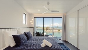 Une chambre moderne avec un grand lit, un ventilateur de plafond et une vue sur une étendue d’eau grâce à de grandes fenêtres.