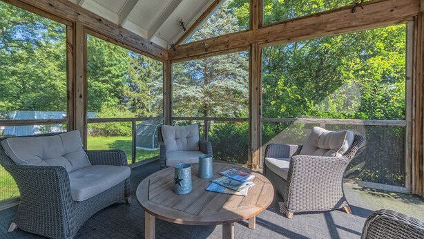 A covered outdoor seating area with wicker furniture and a round table, surrounded by large windows offering a view of a green landscape.