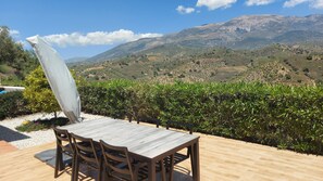 An outdoor seating area with a table and chairs, a white umbrella, and a view of a mountainous landscape.