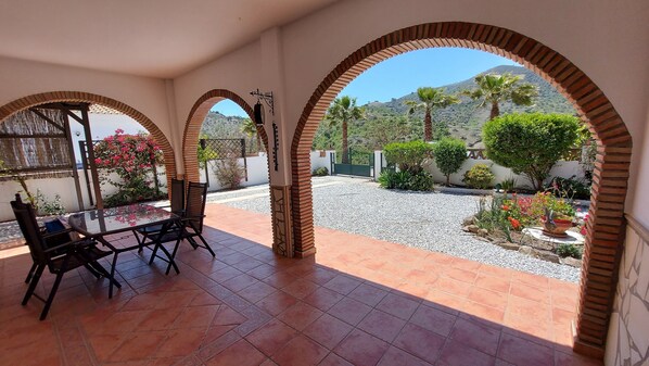 A covered patio with a table and chairs, surrounded by arches and a view of a landscaped garden and distant hills.