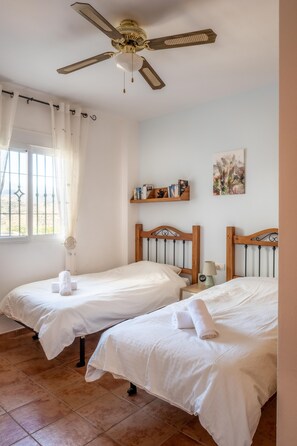 A room with two single beds, a ceiling fan, a window with curtains, a shelf with books, and a painting on the wall.
