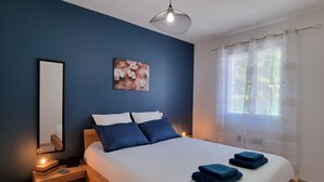 A bedroom with a wooden bed frame, white bedding, and blue pillows. There is a bedside table with a lamp, a mirror, and a small basket. A painting hangs on the wall above the bed.