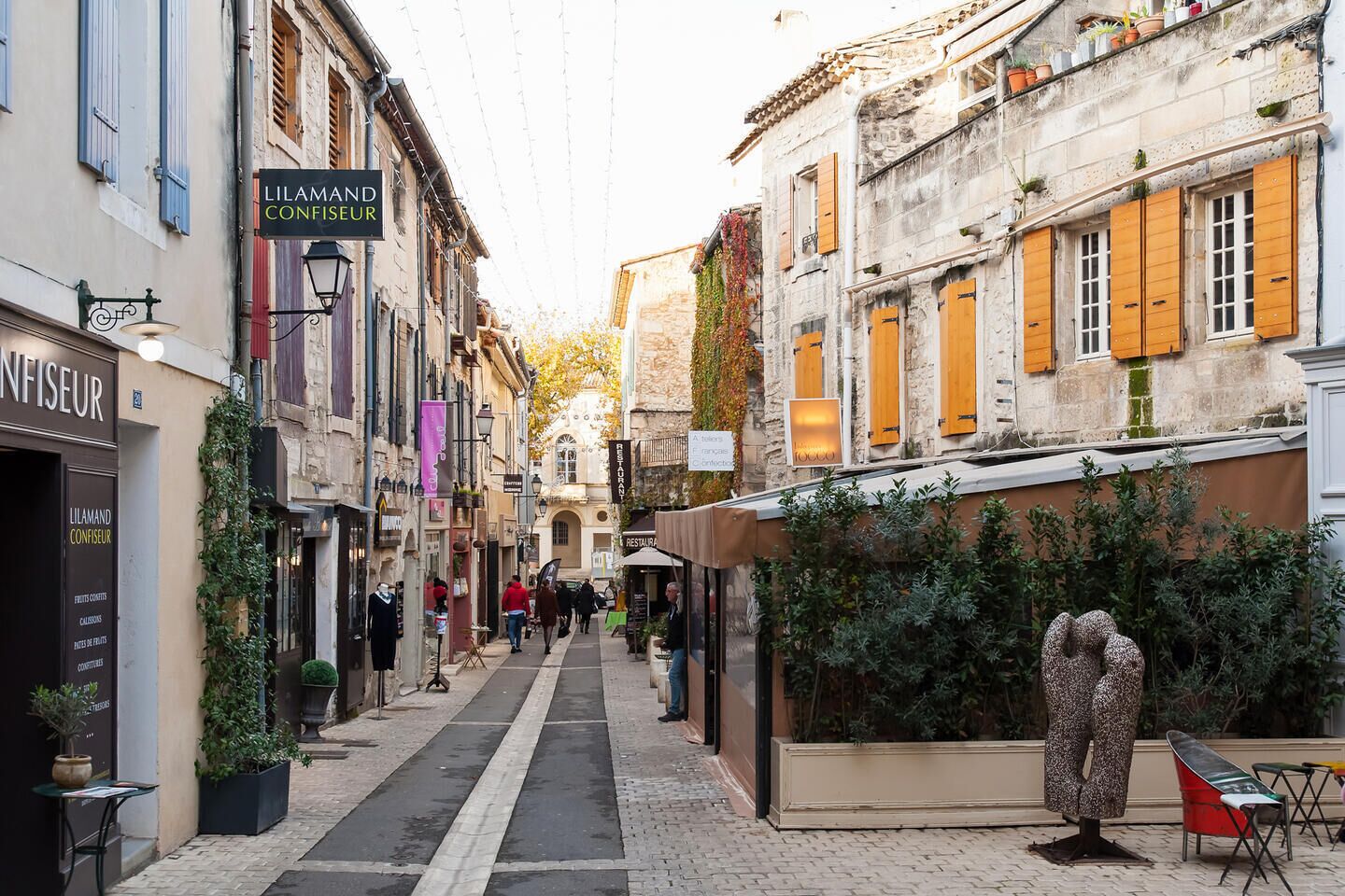A narrow street lined with historic buildings, a café named 'LILAMAND CONFISEUR', and a statue in the foreground.