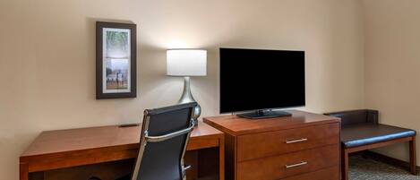 A hotel room with a wooden desk, a black office chair, a television, a lamp, a framed picture on the wall, and a bench.