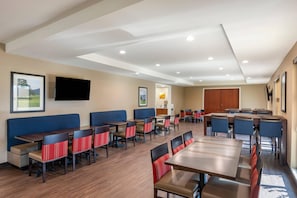 A hotel dining area with multiple tables and chairs, a flat-screen TV mounted on the wall, and framed pictures on the walls.