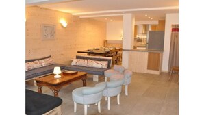 A living room with a sofa, coffee table, and ottomans. There is a dining area with a table and chairs. The room has wooden walls and a kitchen area with a refrigerator and cabinets. - No. 6, maximum 14 people (Vars)