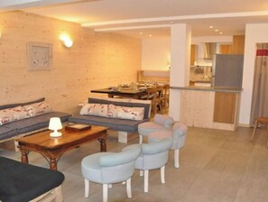 Un salon comprenant un canapé, une table basse et des poufs. On y trouve également un coin repas avec une table et des chaises. Les murs de la pièce sont en bois, et il y a un coin cuisine avec un réfrigérateur et des placards.